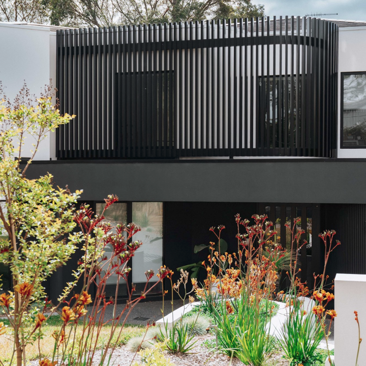 Modern two‑story house with black vertical louvers on the upper floor, large windows, and a foreground garden with red-topped flowering plants.