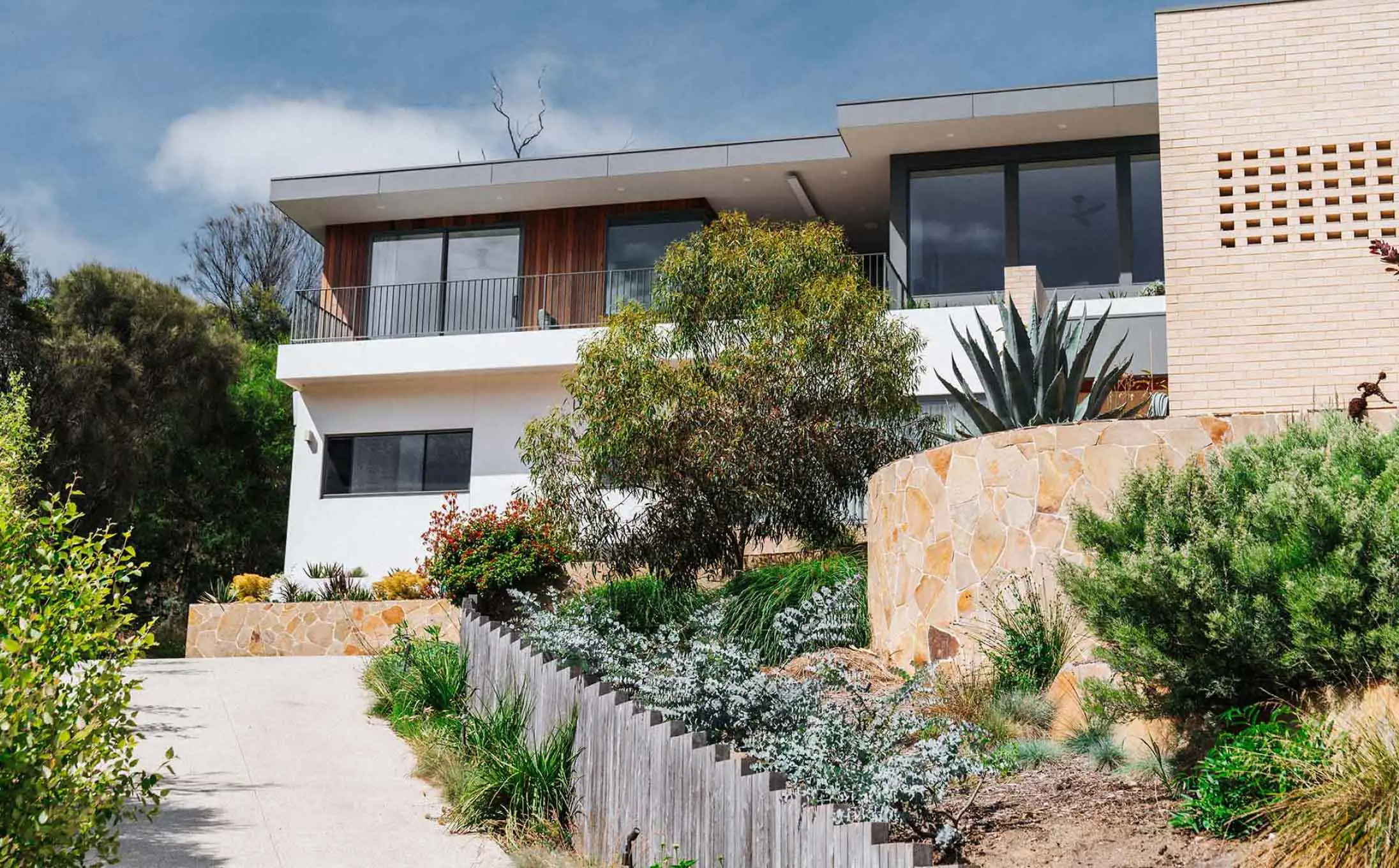 Modern two-story house with a balcony, large glass doors, and a stone retaining wall amid drought-tolerant plants on a hillside.