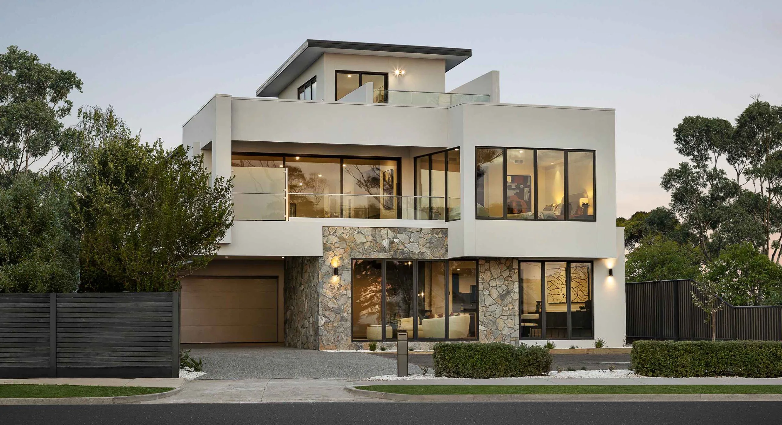 Modern white multi-story house with large glass windows and stone facade, surrounded by trees and a black fence.