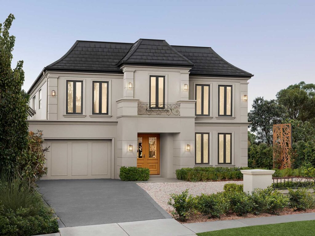Two-story beige townhouse with a black tile roof, central balcony, and wooden front door, surrounded by manicured shrubs and a gravel garden edge.