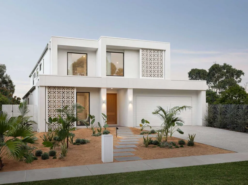 Front view of a modern white two-story house with large windows, decorative lattice panels, and a double garage amid desert landscaping.