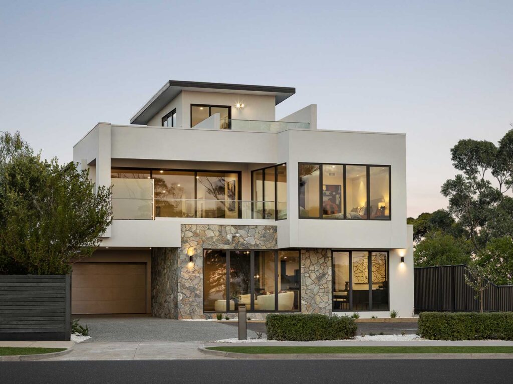 Modern two-story house with white exterior, large glass windows, and a stone accent wall at the ground level.