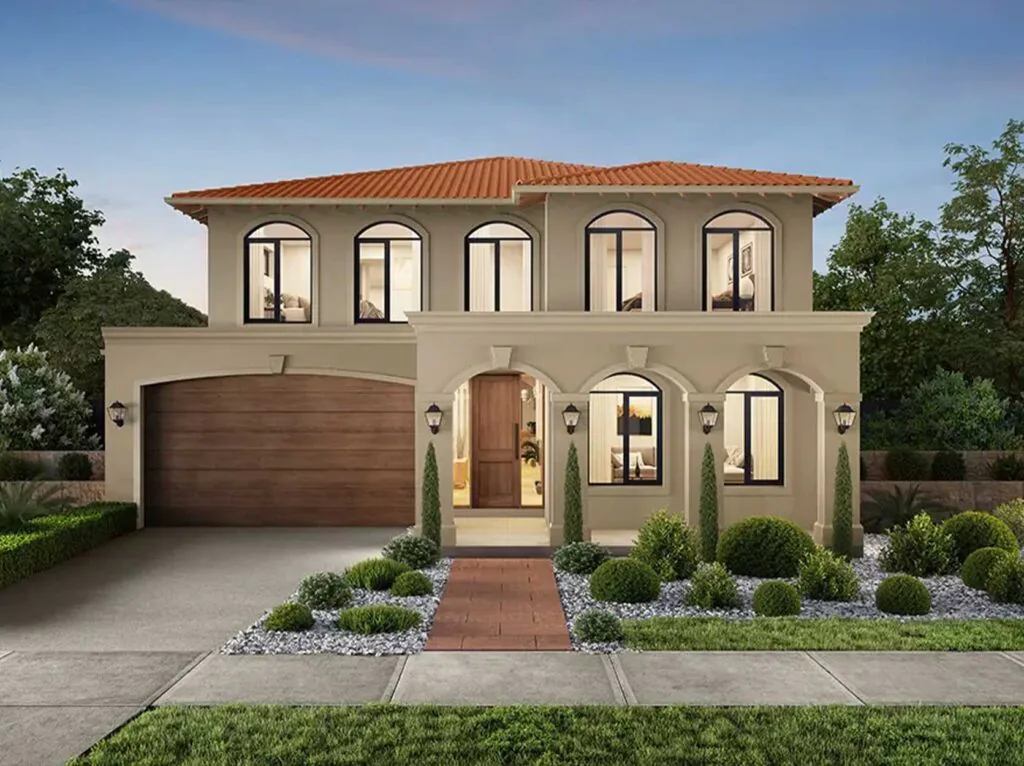 Two-story beige Mediterranean-style house with a red-tiled roof, arched windows, and a wooden front door surrounded by manicured shrubs.