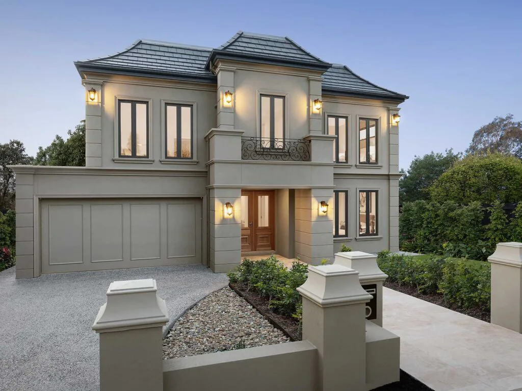 Two-story beige stucco house with a two-car garage, decorative balcony, and warm lanterns on the facade at dusk.