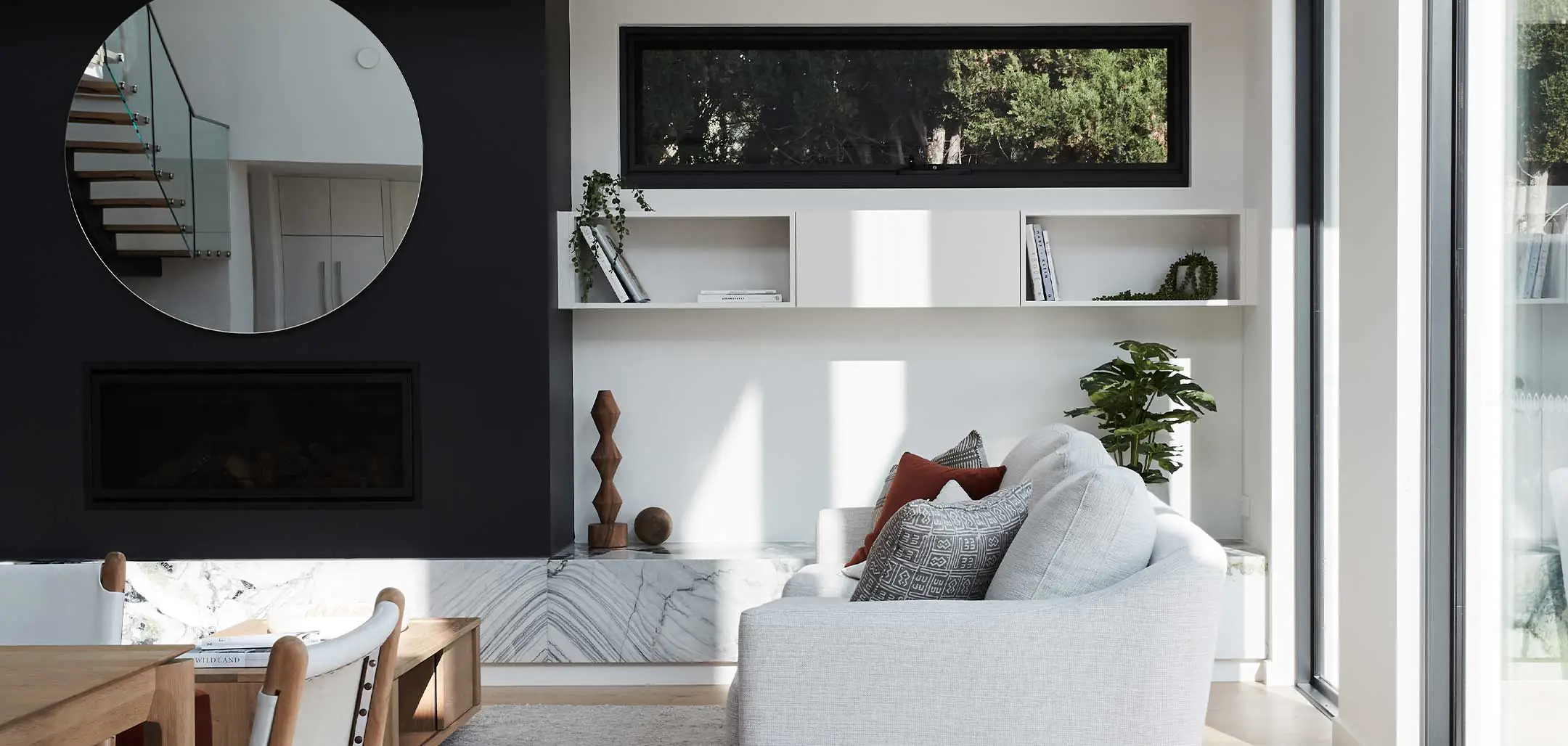 Modern living room with a circular wall mirror, built-in white shelves, and a black fireplace on the left; light gray sofa and greenery by the windows.