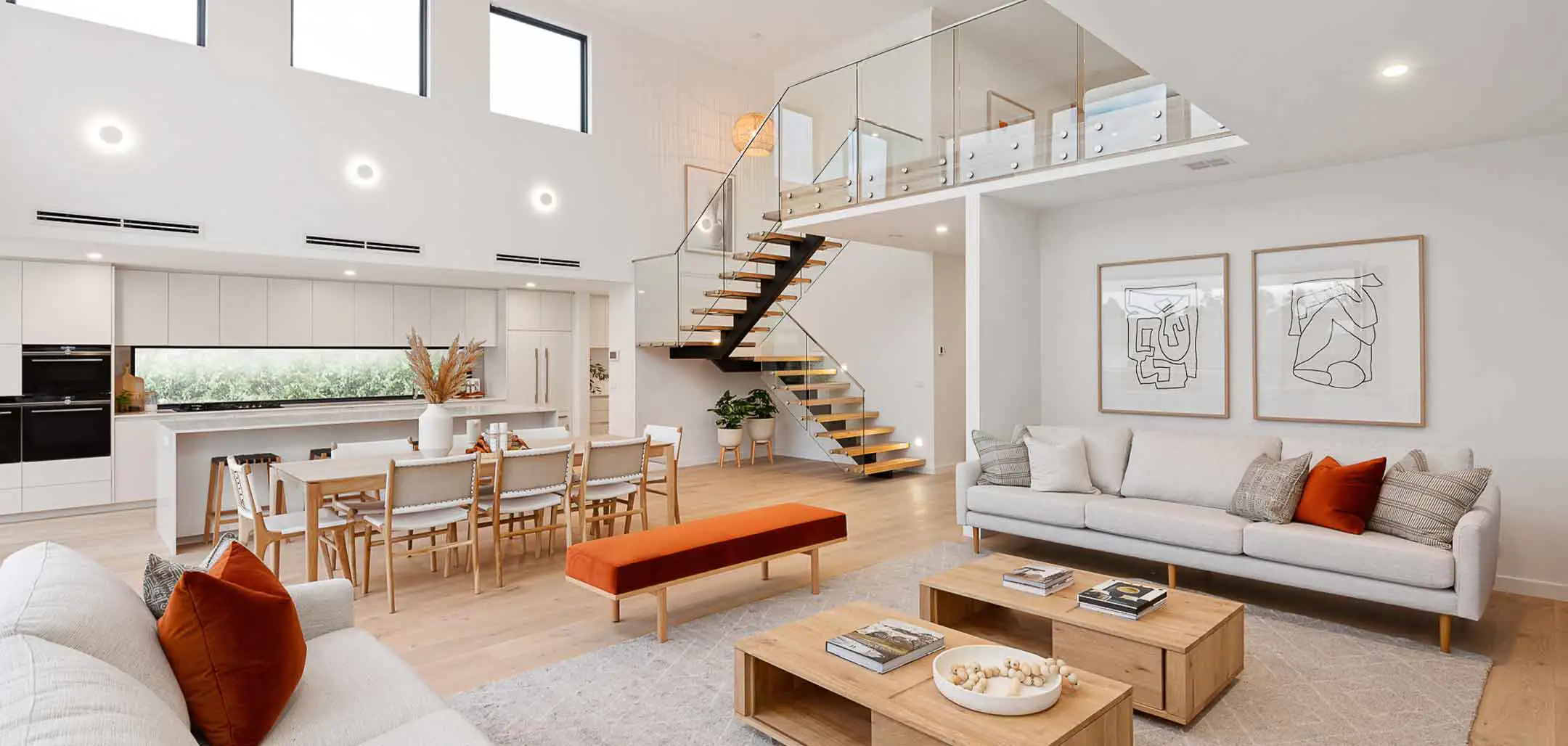 Open-plan living room with staircase to mezzanine, white kitchen, wooden furniture, and abstract framed art on the wall near a glass railing.