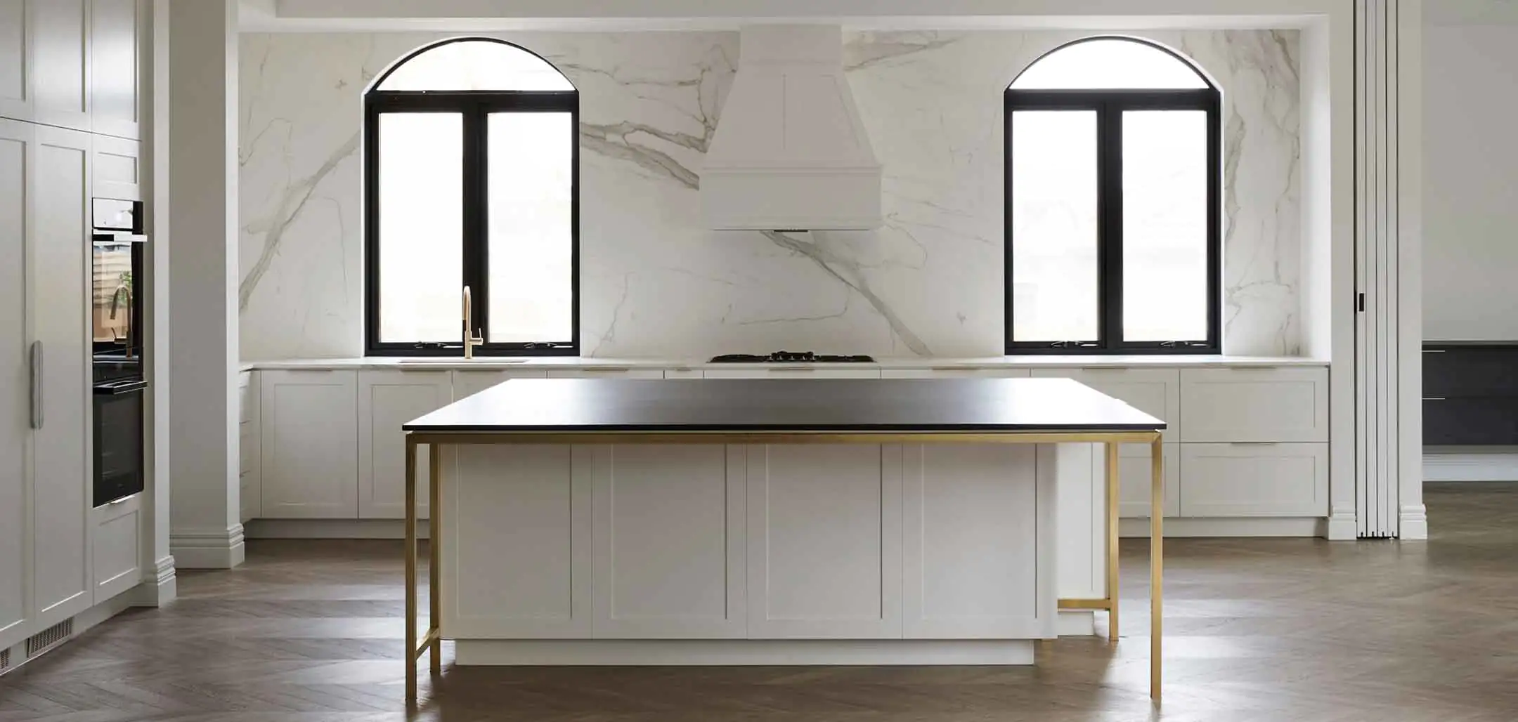 Bright modern kitchen with marble walls, two arched black-framed windows, and a white island with gold legs.