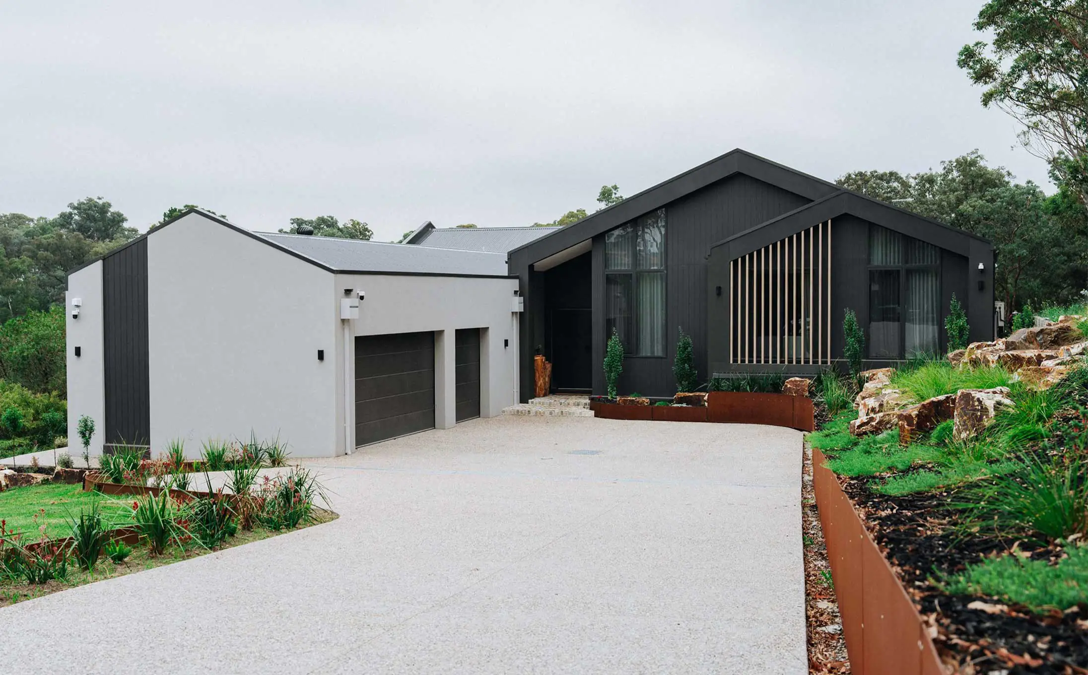 Modern contemporary house with dark gray exterior, large windows, and a two-car garage, set in a landscaped yard along a gravel driveway.