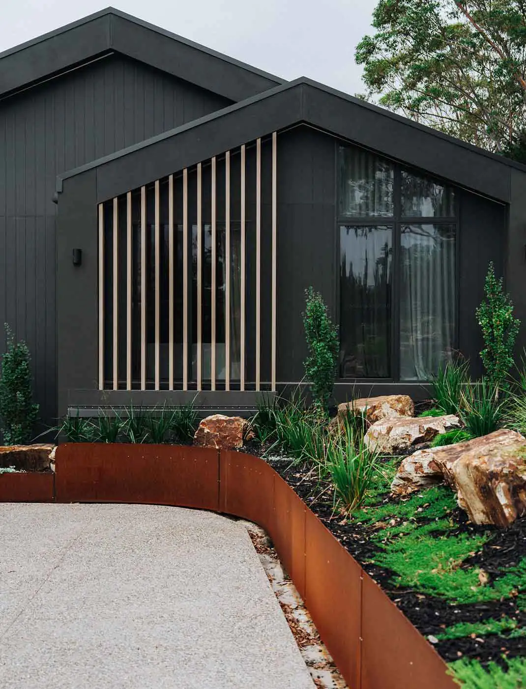 Modern dark gray house exterior with a slatted wooden screen along a large window, surrounded by rocks and greenery.
