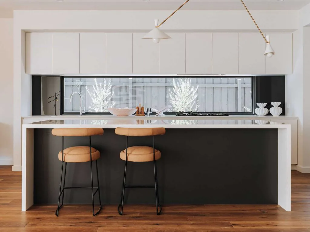 Modern white kitchen with a glossy island, two tan upholstered bar stools, and a large window behind the sink.