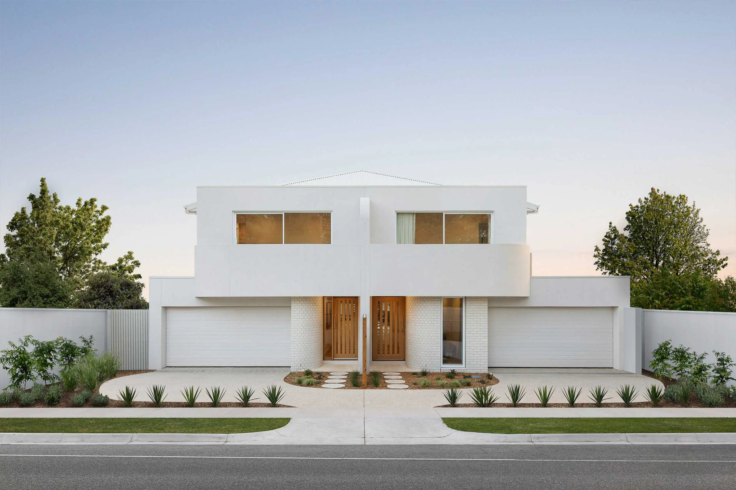 Front view of a modern white two-story house with large windows, a wooden front door, and a manicured drought-tolerant garden lining the walkway.