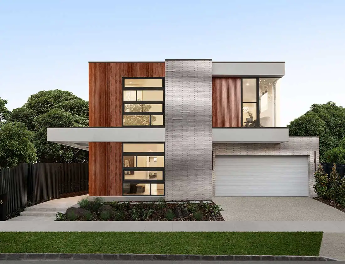 Two-story modern house with gray brick central column, wood siding, large windows, and a double garage to the right, sunny setting.