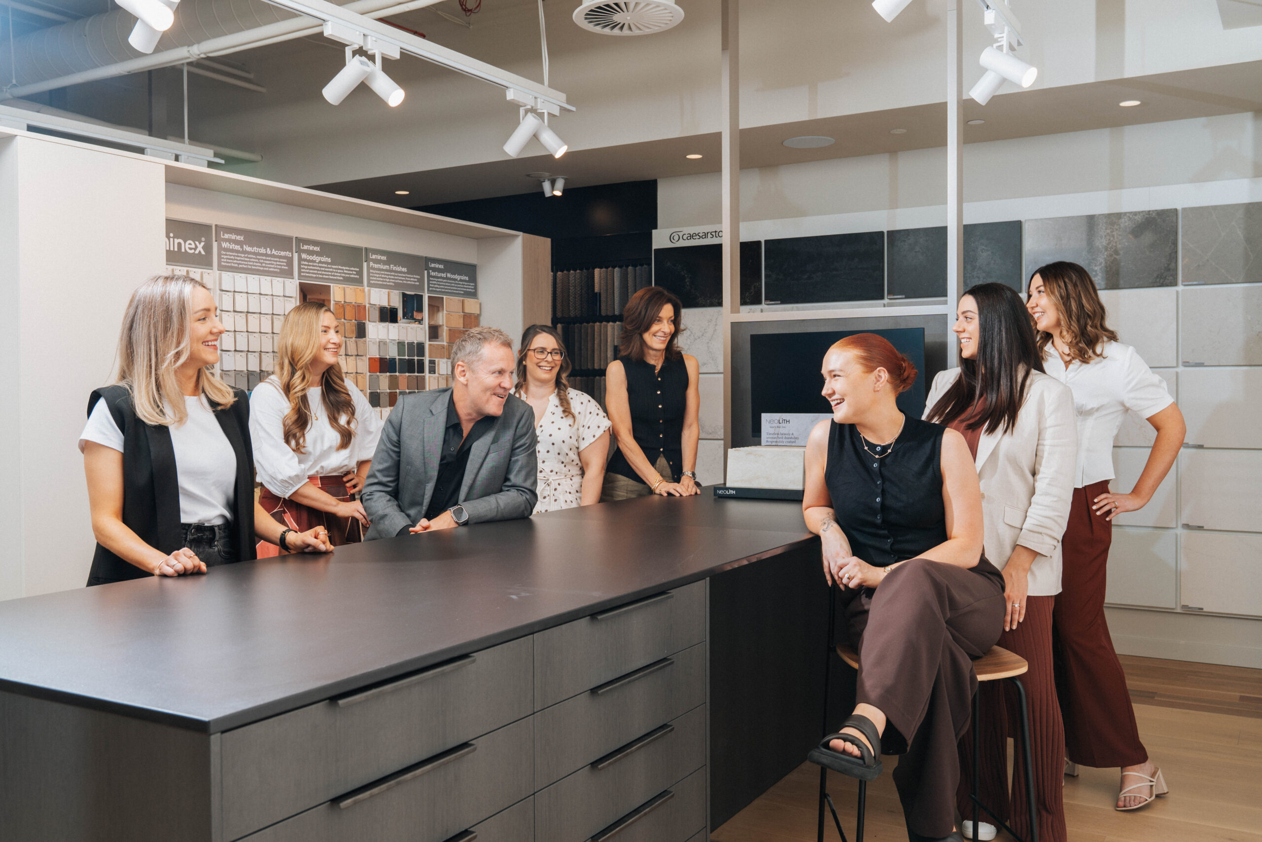 Group of professionals gathered around a dark reception counter in a showroom, smiling and chatting near fabric swatches and tile samples.