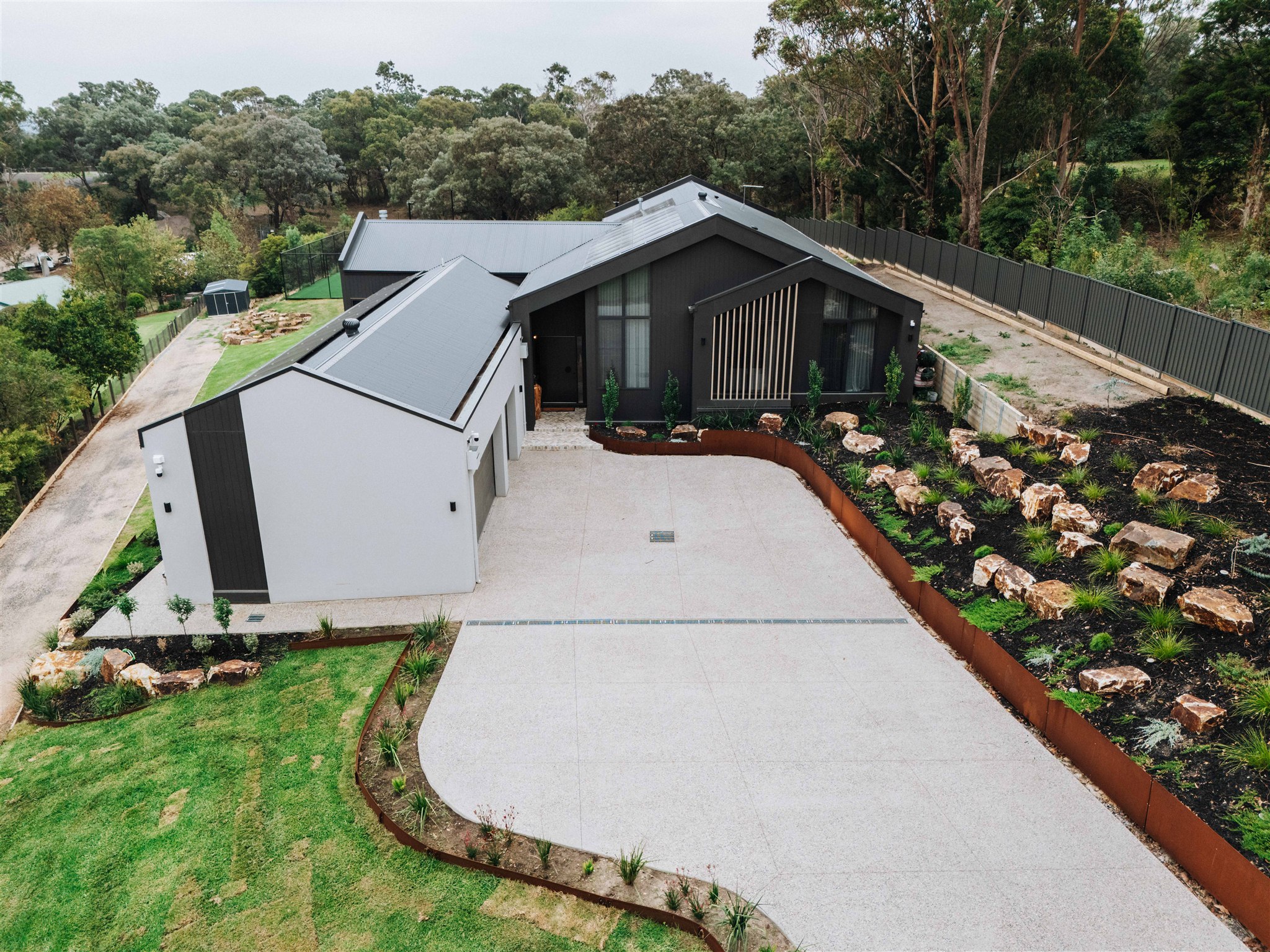 Aerial view of a modern dark-gray house with a metal roof, concrete driveway, and landscaped rock garden. (informative)