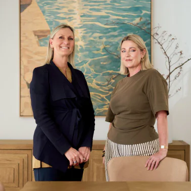 Two women stand side by side in a modern office, smiling, with a blue-green abstract painting behind them.