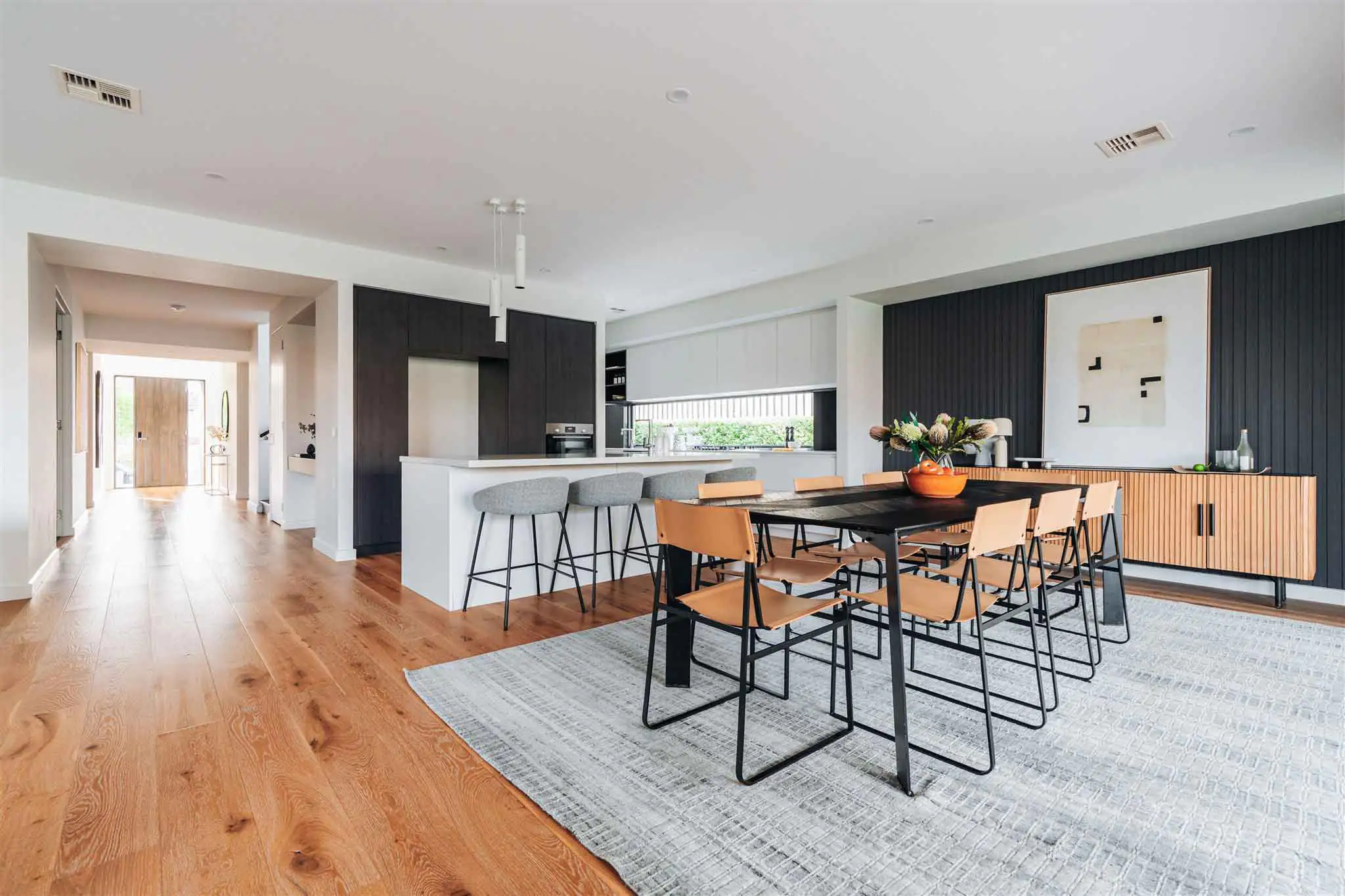 Open-plan kitchen and dining area with a long black-framed dining table and tan chairs under pendant lights.