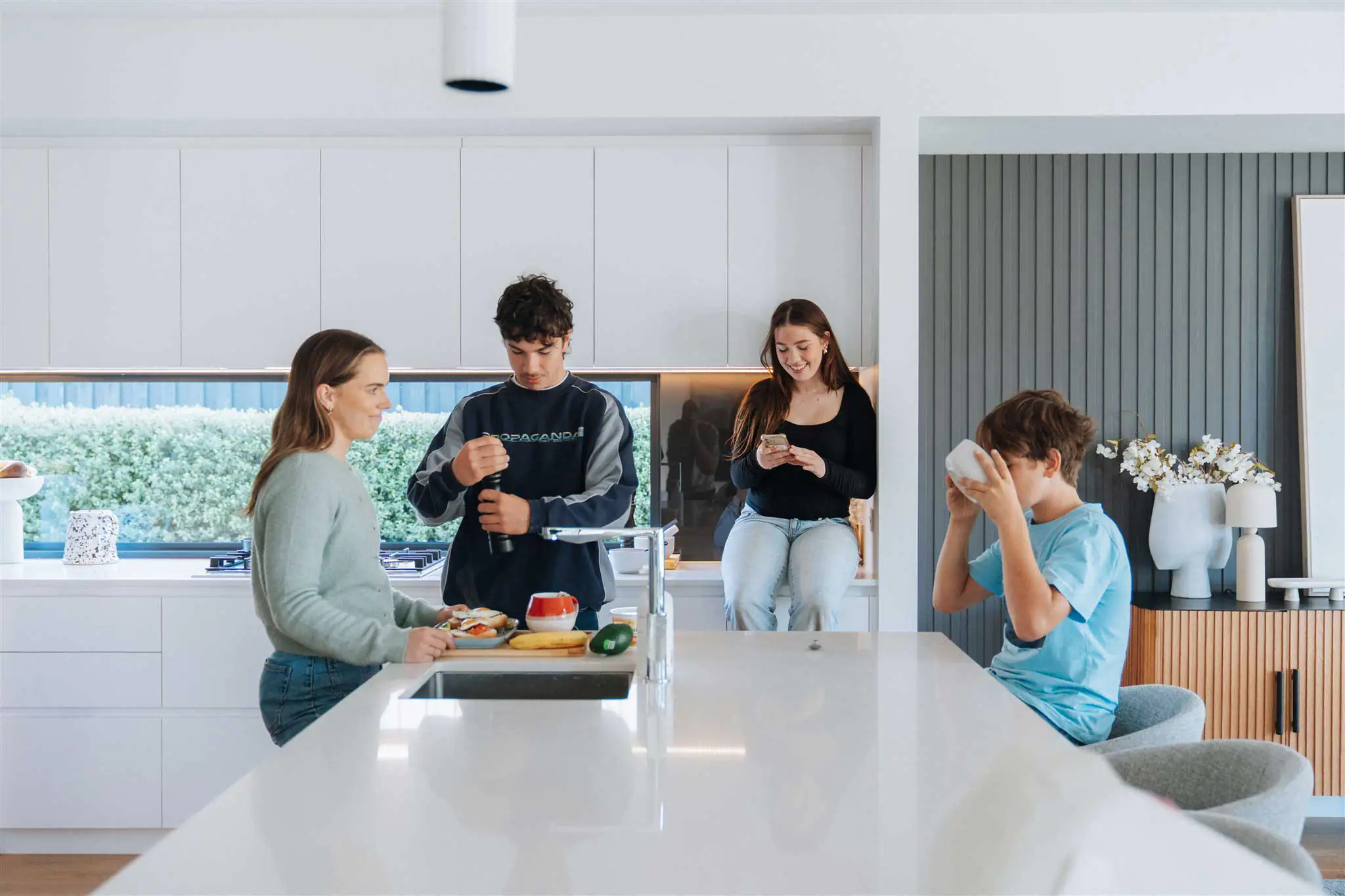 Four friends socialize in a bright, modern kitchen around a large white island, with food prep underway in the foreground and a person on a phone in the background.
