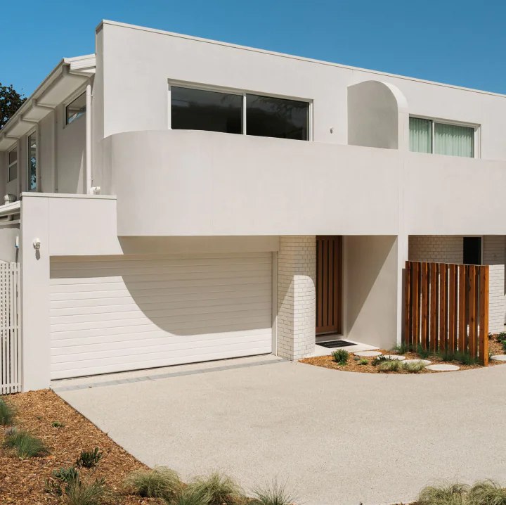 Modern white two-story house with a large garage door, wooden entry door, and low landscaping under a clear blue sky