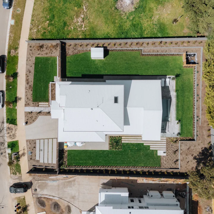 Aerial view of a white-roofed house with a manicured green lawn, stone paths, and surrounding low walls; driveway and street visible on the left.