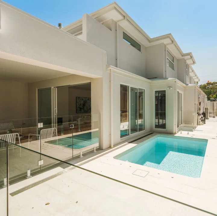 Modern two-story white house with a glass-railed patio and a rectangular turquoise pool in the sunny backyard.