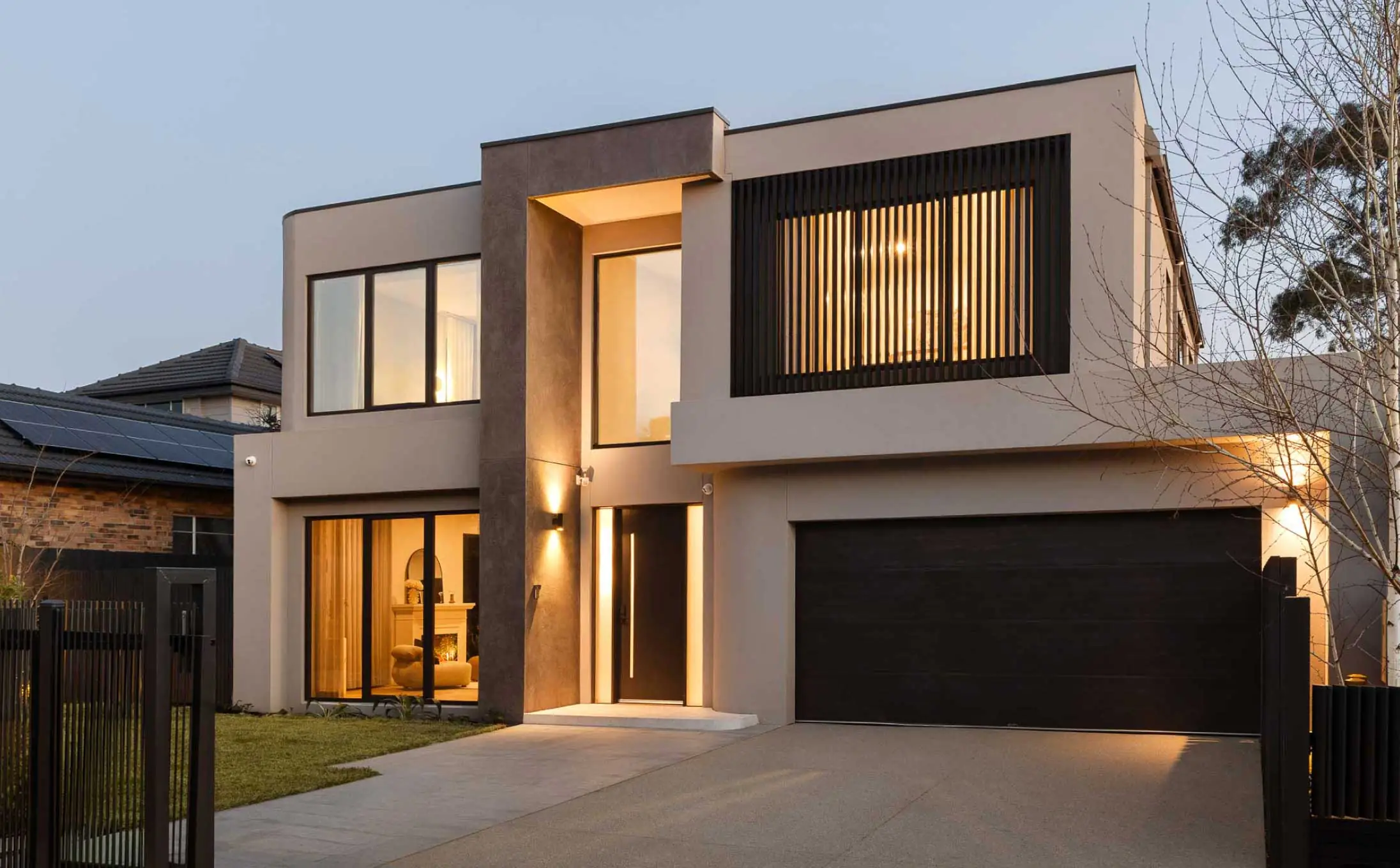 Front view of a modern two-story house with large glass windows, warm interior lighting, and a dark garage door at dusk.
