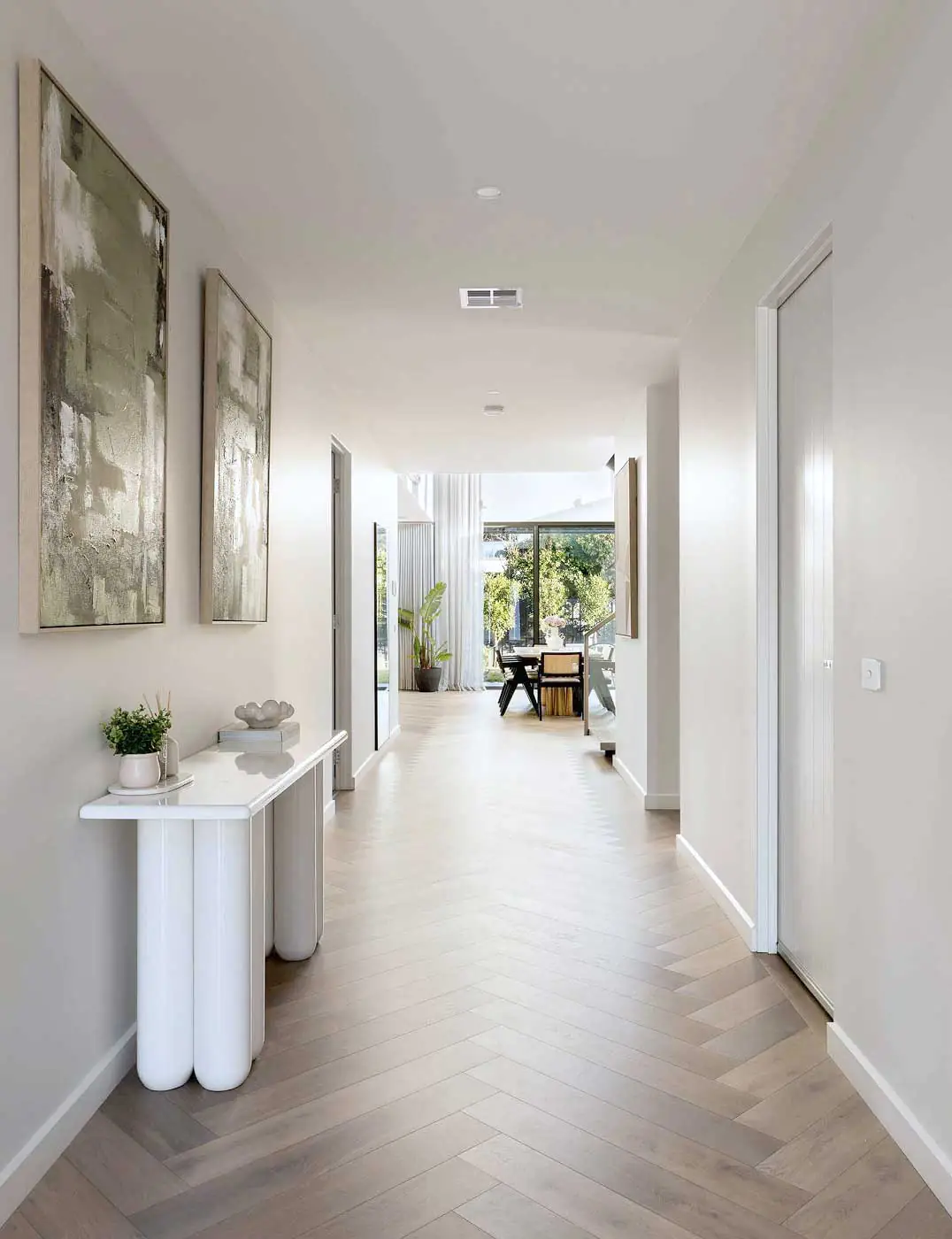 Bright modern hallway with light wood chevron flooring, abstract left-wall art, and a white console table with plants leading to a sunlit living area.