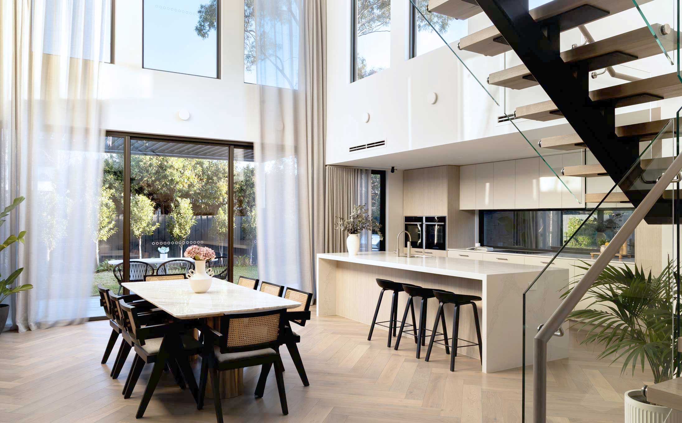 Open-plan modern kitchen and dining area with a long marble table, black chairs, large windows, and a glass-railed staircase overlooking a garden outside.