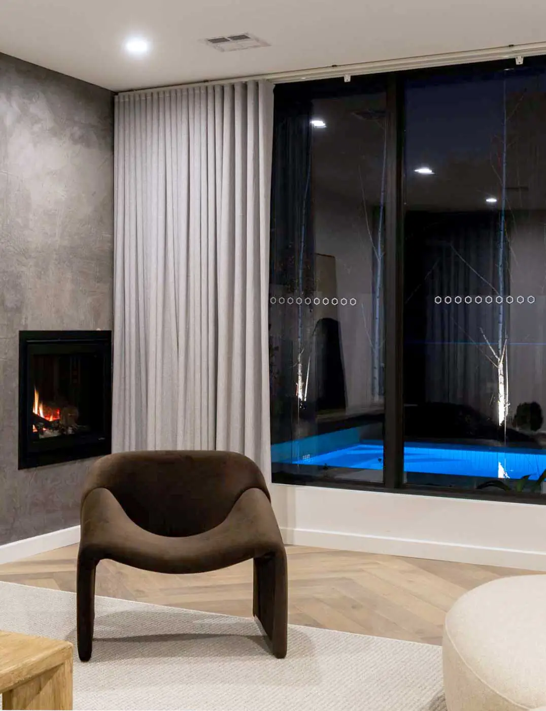 Modern living room at night with a built-in fireplace, curved brown lounge chair, light gray curtains, and a glass wall overlooking a blue-lit pool.