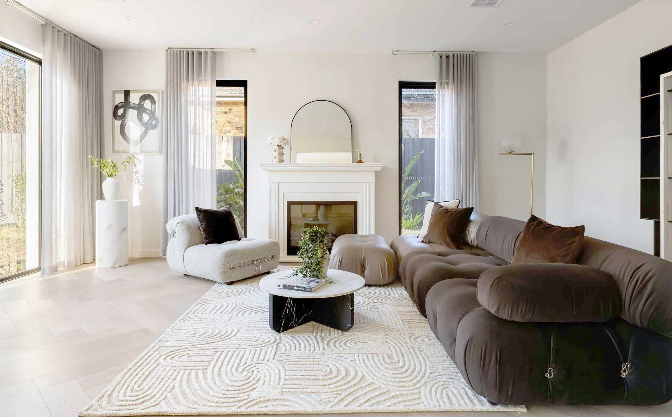Bright living room with a white fireplace, arched mirror, and neutral seating under sheer curtains.