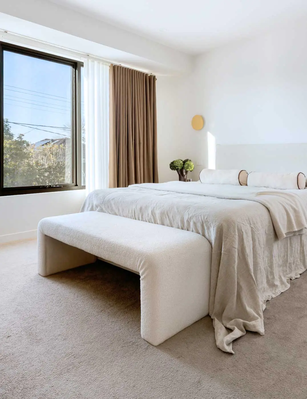 Bright minimalist bedroom with a white bed, cream bedding, a curved bench at the foot, and beige curtains by a large window.